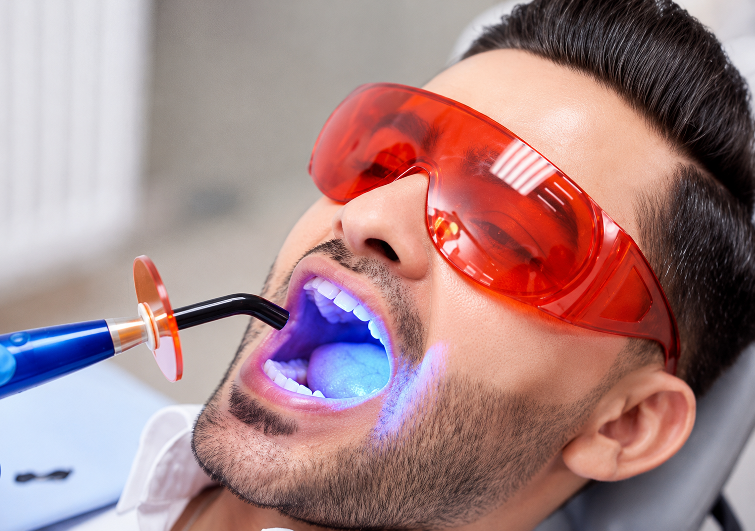 Dental patient wearing protective red glasses during a laser dentistry treatment, with a blue curing light visible at the mouth.